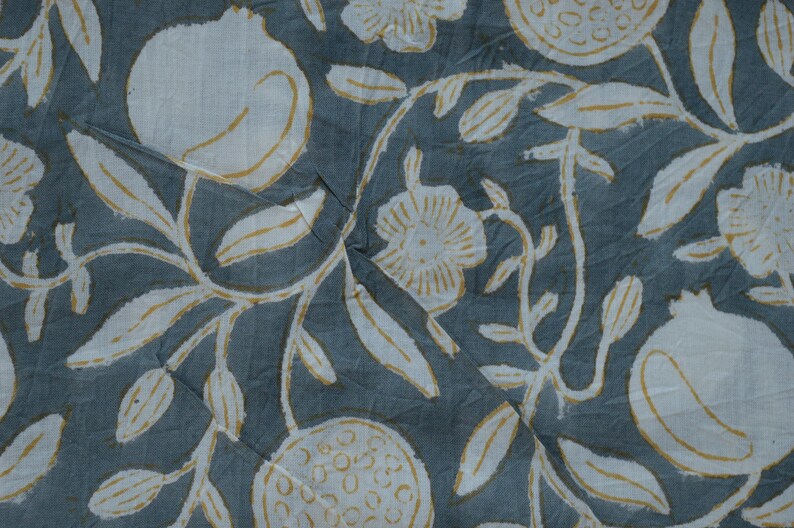 May include: Close-up of a fabric with a repeating pattern of white pomegranates, flowers, and leaves with yellow accents on a muted blue-gray background. The design has a hand-drawn, block-print style.