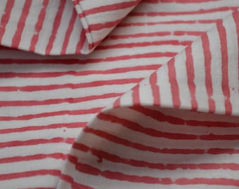 May include: Close-up of white fabric with a repeating pattern of thick, uneven coral-colored stripes. The fabric is folded, revealing the pattern from different angles. The material appears to be cotton or a similar textile.