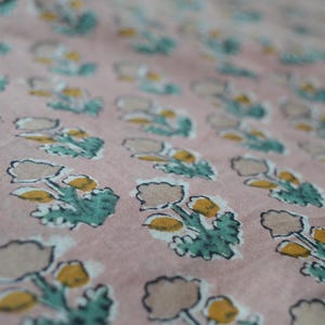 May include: Close-up of a fabric with a repeating floral pattern. The flowers have tan petals and yellow centers, with teal green leaves. The background is a muted pink color.