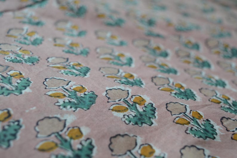 May include: Close-up of a fabric with a repeating floral pattern. The flowers have tan petals and yellow centers, with teal green leaves. The background is a muted pink color.