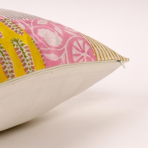 Op de afbeelding: Een decoratief kussen met een witte basis en een kleurrijk patchwork-ontwerp. Het patchwork bevat geel met groene accenten, roze bloempatronen en gouden en zwarte strepen. Een ritssluiting is aan de zijkant zichtbaar.