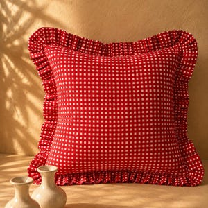 May include: A square throw pillow featuring a red and white gingham check pattern. The pillow is finished with a ruffled border made of the same red and white checkered fabric, creating a decorative edge around the entire perimeter of the cushion.
