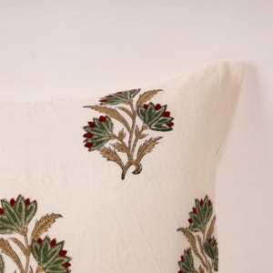 May include: Off-white pillow with a floral pattern. The design features green leaves, red flower buds, and tan stems. The pillow has a soft, textured appearance, suggesting a comfortable home decor item.