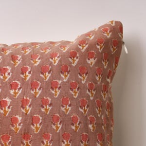 Könnte beinhalten: Ein quadratisches Zierkissen mit einem sich wiederholenden Blumenmuster. Die Blumen sind in Rot-, Orange- und Gelbtönen gehalten, vor einem gedämpften rosa Hintergrund. Ein weißer Reißverschluss ist an der Seite sichtbar.