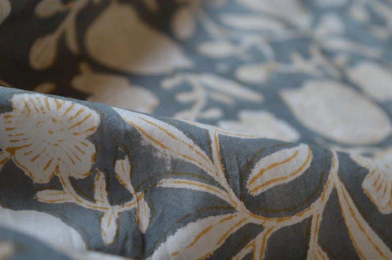 May include: Close-up of a fabric with a floral pattern. The fabric is a muted blue-gray color with white and yellow floral designs. The fabric is folded, showing the texture and detail of the print.