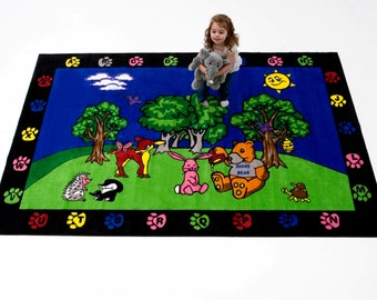 May include: A colorful children's rug featuring a playful scene with cartoon animals, trees, and a sunny sky. The rug has a black border with paw prints and letters. A young child is standing on the rug holding a stuffed elephant.