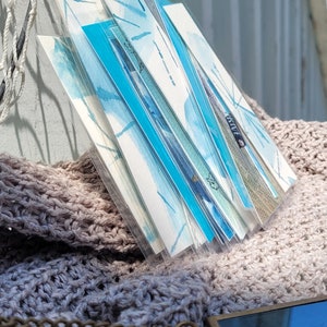 May include: A stack of watercolor art prints in clear plastic sleeves, featuring abstract blue and white designs. The prints are displayed on a textured, light brown surface, with a small, rectangular pendant in the foreground.