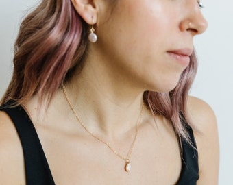May include: A gold necklace with a small, round, white pendant. Matching gold earrings with similar white, round stones. The jewelry is worn with a black tank top.