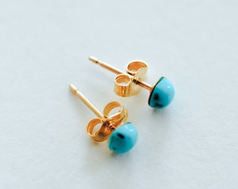 May include: A pair of gold-colored stud earrings with turquoise-colored, half-sphere stones. The earrings are displayed on a white surface, showcasing their simple yet elegant design. The posts and backs are visible, highlighting the jewelry's construction.