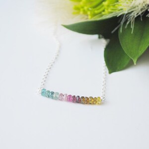 May include: A delicate necklace featuring a strand of colorful tourmaline beads in shades of blue, pink, and yellow. The gemstones are strung on a fine silver chain. The necklace is displayed on a white surface with green foliage in the background.