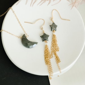 May include: A gold necklace with a crescent moon pendant and a pair of star earrings with gold chain tassels. The jewelry is displayed on a white plate against a black and white background.