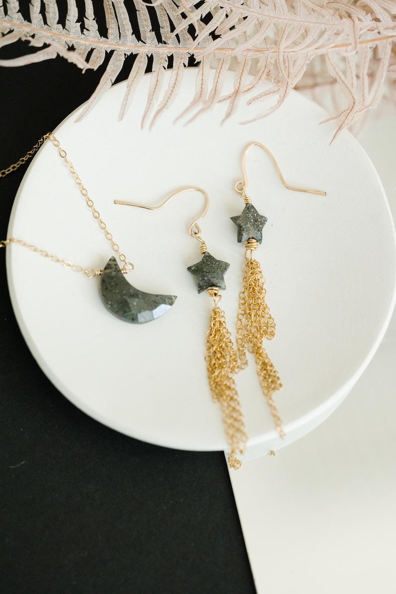 May include: A gold necklace with a crescent moon pendant and a pair of star earrings with gold chain tassels. The jewelry is displayed on a white plate against a black and white background.