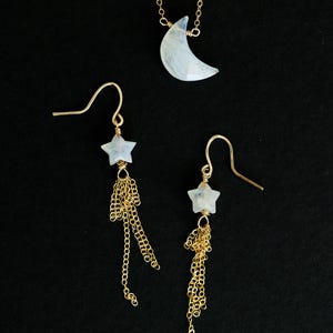 Puede incluir: Un collar de oro con un colgante de media luna y un par de pendientes de oro. Los pendientes presentan piedras preciosas en forma de estrella y cadenas de oro colgantes. La joyería está sobre un fondo negro.