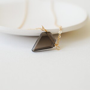 May include: A faceted, smoky quartz pendant necklace. The dark gray crystal is suspended from a delicate gold-colored chain. The necklace is displayed on a white surface, creating a minimalist aesthetic.