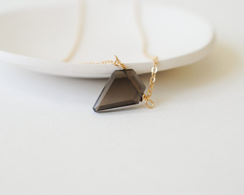 May include: A faceted, smoky quartz pendant necklace. The dark gray crystal is suspended from a delicate gold-colored chain. The necklace is displayed on a white surface, creating a minimalist aesthetic.