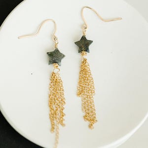 May include: A pair of gold-colored dangle earrings. Each earring features a dark gray star-shaped bead and multiple strands of gold-colored chain. The earrings are displayed on a white surface.