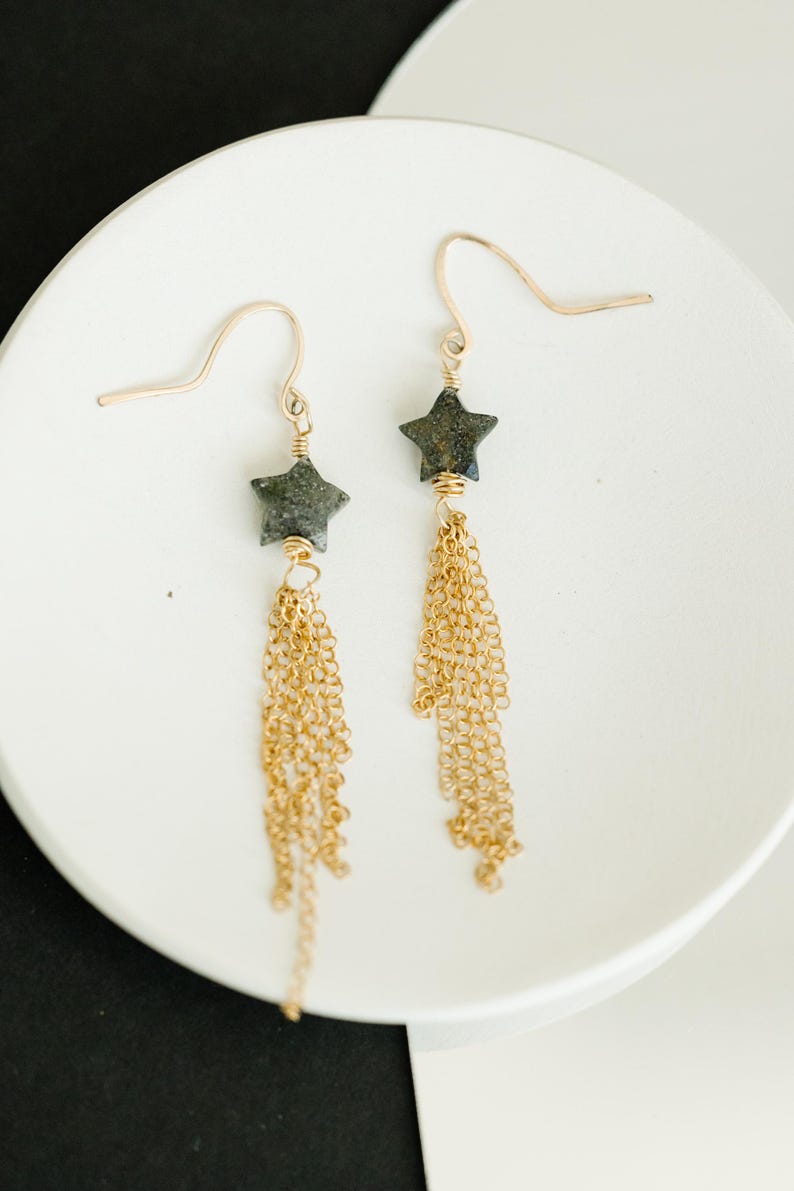 May include: A pair of gold-colored dangle earrings. Each earring features a dark gray star-shaped bead and multiple strands of gold-colored chain. The earrings are displayed on a white surface.