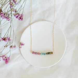 May include: A delicate gold necklace featuring a row of colorful tourmaline beads. The beads transition from pink to green, creating a gradient effect. The necklace is displayed on a white ceramic dish, with pink flowers in the background.