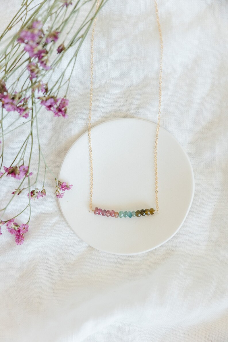 May include: A delicate gold necklace featuring a row of colorful tourmaline beads. The beads transition from pink to green, creating a gradient effect. The necklace is displayed on a white ceramic dish, with pink flowers in the background.