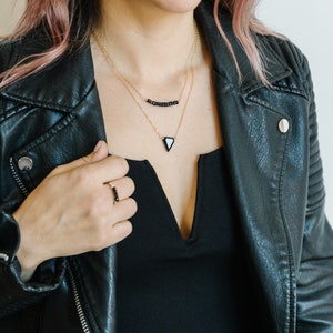 May include: A woman wearing a black leather jacket, a black top, and gold necklaces. The necklaces feature black beads and a black triangle pendant. A matching black beaded ring is on the woman's finger.
