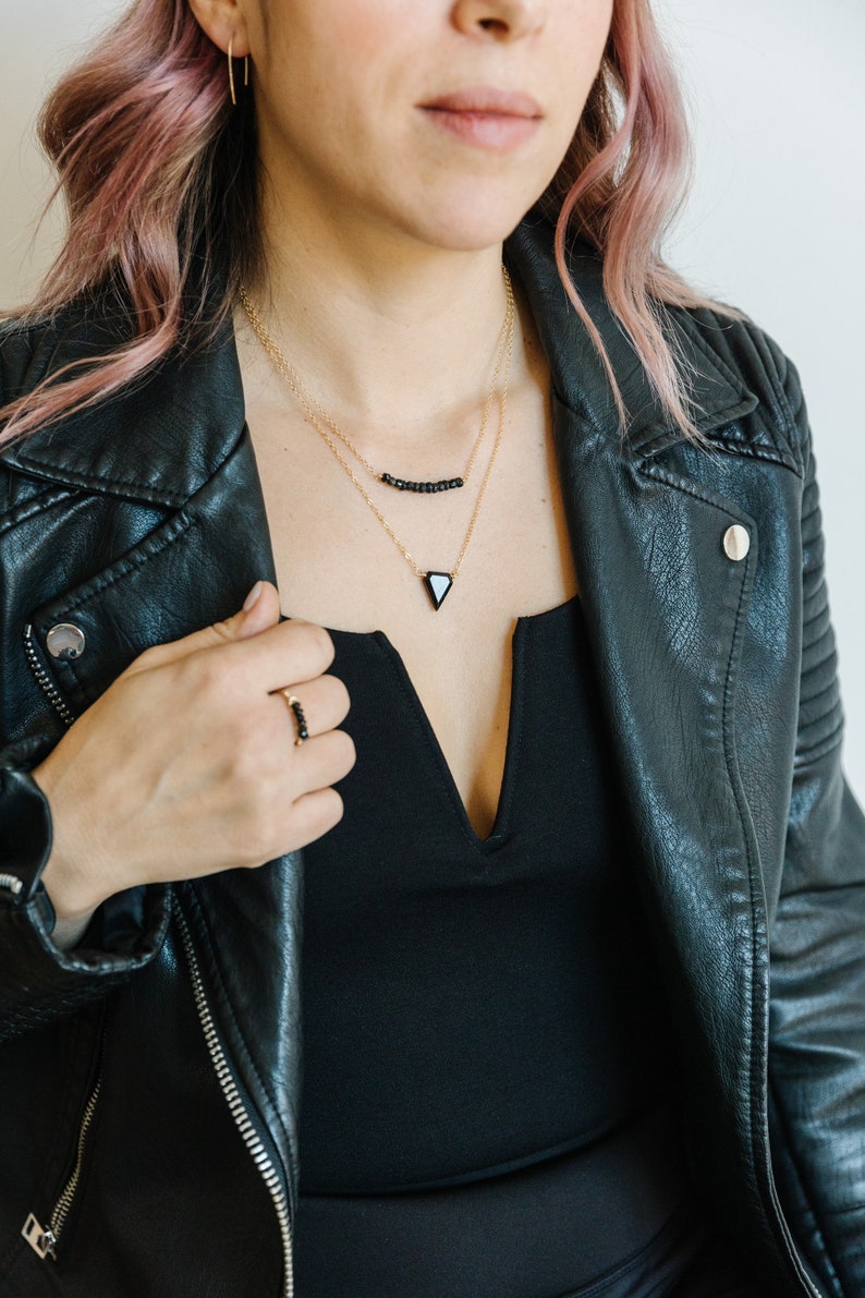 May include: A woman wearing a black leather jacket, a black top, and gold necklaces. The necklaces feature black beads and a black triangle pendant. A matching black beaded ring is on the woman's finger.