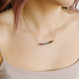 May include: A delicate gold necklace featuring a row of colorful tourmaline beads. The beads transition in color from pink to green and brown. The necklace is worn on a person's neck, against a neutral-toned top.