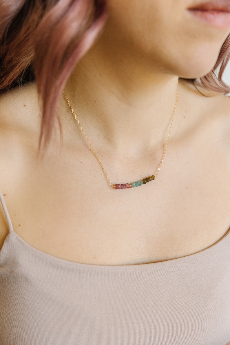 May include: A delicate gold necklace featuring a row of colorful tourmaline beads. The beads transition in color from pink to green and brown. The necklace is worn on a person's neck, against a neutral-toned top.