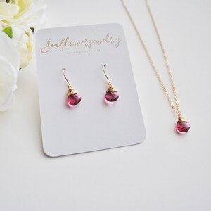 Puede incluir: Un conjunto de joyas con un collar y pendientes dorados, cada uno con una piedra preciosa rosa en forma de l&aacute;grima. Los pendientes son de tipo gancho. El collar y los pendientes se muestran en una tarjeta blanca con texto.