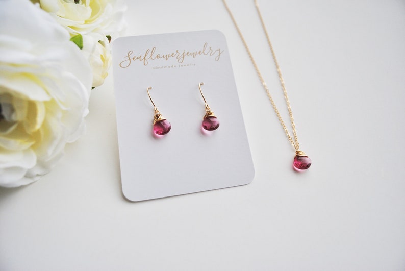 Puede incluir: Un conjunto de joyas con un collar y pendientes dorados, cada uno con una piedra preciosa rosa en forma de l&aacute;grima. Los pendientes son de tipo gancho. El collar y los pendientes se muestran en una tarjeta blanca con texto.
