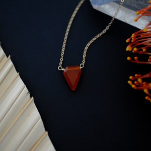 May include: A gold chain necklace with a triangular, orange-red gemstone pendant. The pendant is faceted and catches the light. The necklace is displayed on a dark background with a dried palm leaf and orange flower accents.