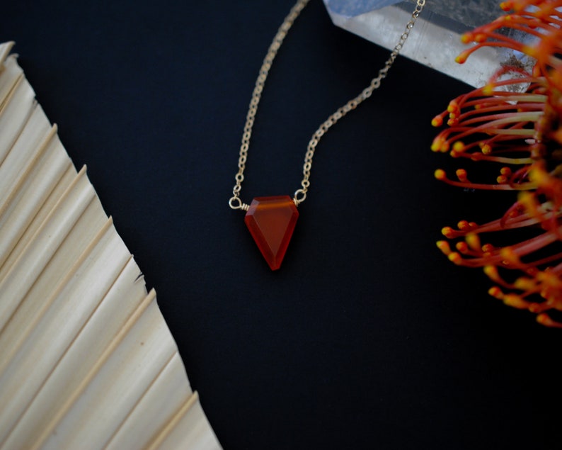 May include: A gold chain necklace with a triangular, orange-red gemstone pendant. The pendant is faceted and catches the light. The necklace is displayed on a dark background with a dried palm leaf and orange flower accents.