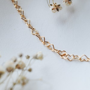May include: A delicate gold-colored necklace featuring a chain of interconnected heart-shaped links. The necklace is displayed on a white surface, with dried white flowers in the background. The jewelry is a romantic accessory.
