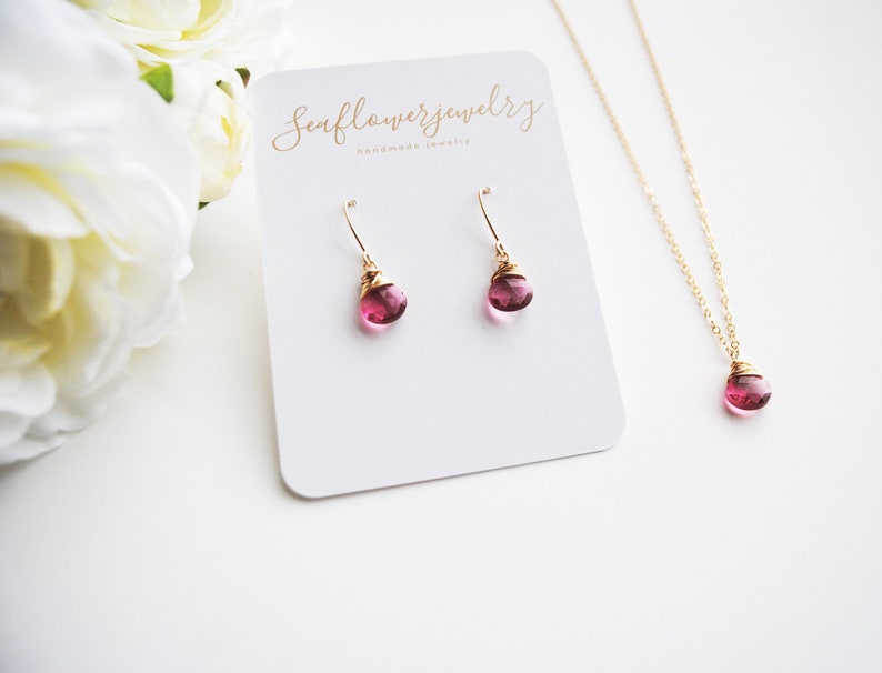 Puede incluir: Un conjunto de collar y pendientes dorados con un peque&ntilde;o colgante redondo de piedra rosa y pendientes a juego. Las joyas se exhiben en una tarjeta blanca con el texto "Seaflowerjewelry handmade jewelry".