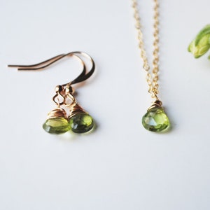 May include: A set of jewelry featuring a pair of gold-toned earrings and a necklace. Each piece has a teardrop-shaped, light green gemstone. The earrings have a hook design, and the necklace has a delicate chain.