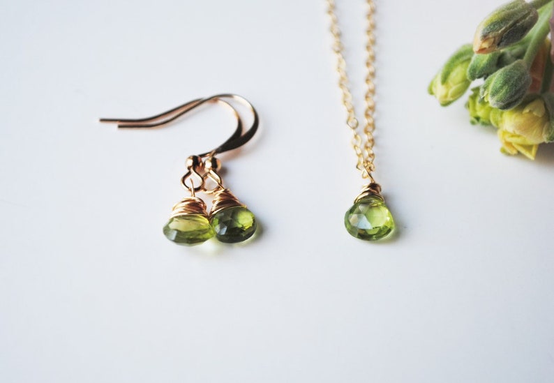 May include: A set of jewelry featuring a pair of gold-toned earrings and a necklace. Each piece has a teardrop-shaped, light green gemstone. The earrings have a hook design, and the necklace has a delicate chain.