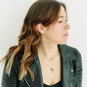 May include: A woman wearing gold necklaces and earrings with dark gray stone pendants. She is wearing a black leather jacket over a black top. Her hair is brown with auburn highlights.