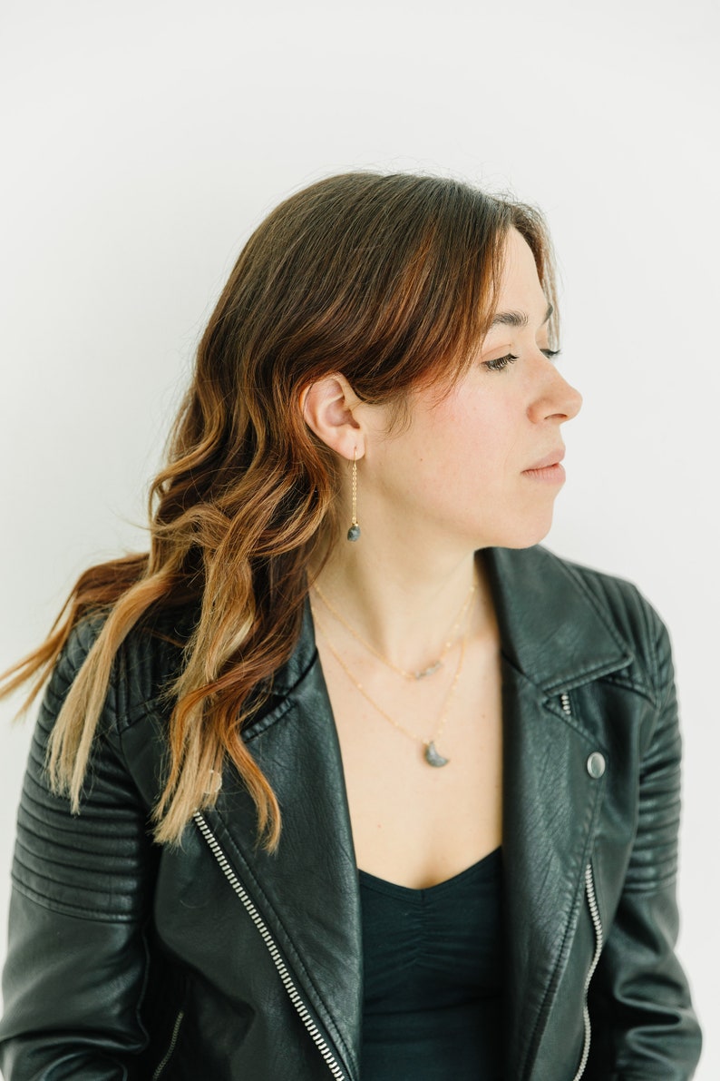 May include: A woman wearing gold necklaces and earrings with dark gray stone pendants. She is wearing a black leather jacket over a black top. Her hair is brown with auburn highlights.