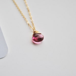 Puede incluir: Un collar de cadena dorada con un colgante de piedra preciosa rosa facetada en forma de l&aacute;grima. El colgante est&aacute; envuelto en alambre y cuelga de una delicada cadena. El fondo es blanco.