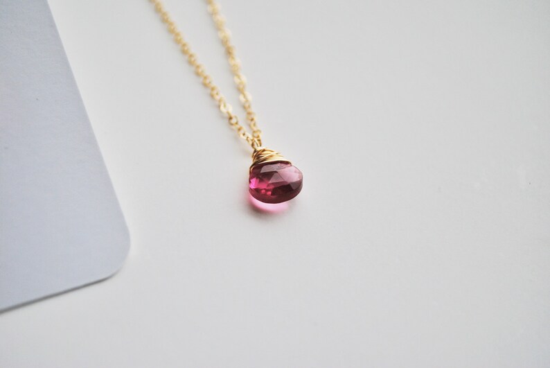 Puede incluir: Un collar de cadena dorada con un colgante de piedra preciosa rosa facetada en forma de l&aacute;grima. El colgante est&aacute; envuelto en alambre y cuelga de una delicada cadena. El fondo es blanco.