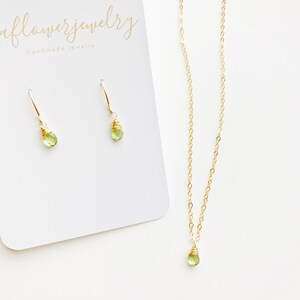 May include: A gold-colored necklace and matching earrings set. The necklace and earrings feature a small, light green gemstone. The earrings are on a white card with the text "aflowerjewelry handmade jewelry".