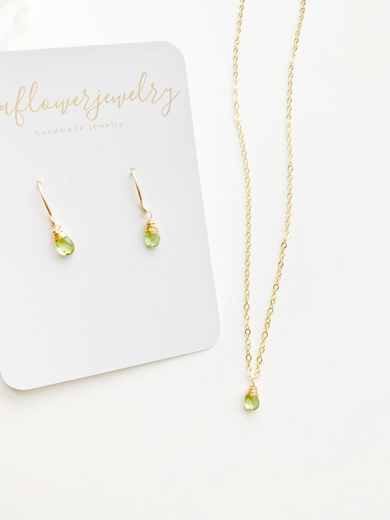 May include: A gold-colored necklace and matching earrings set. The necklace and earrings feature a small, light green gemstone. The earrings are on a white card with the text "aflowerjewelry handmade jewelry".