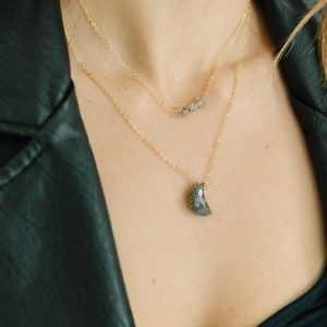 May include: Two layered gold necklaces. The top necklace has a series of small, gray stones. The bottom necklace features a crescent moon-shaped pendant with a gray, speckled pattern. The necklaces are displayed against a black leather jacket.