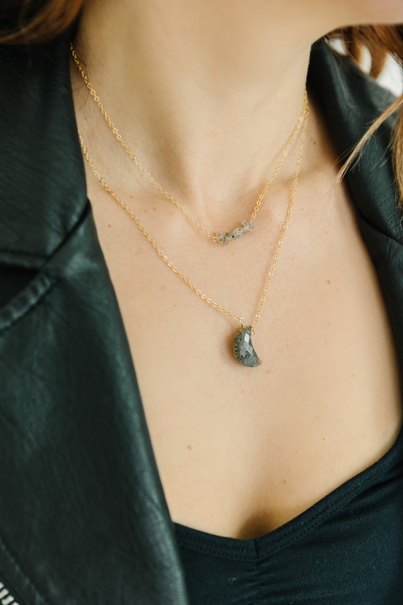 May include: Two layered gold necklaces. The top necklace has a series of small, gray stones. The bottom necklace features a crescent moon-shaped pendant with a gray, speckled pattern. The necklaces are displayed against a black leather jacket.