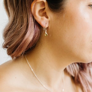 May include: A close-up of a woman wearing a silver necklace and earrings. The earrings are small, with a silver hook and a teardrop-shaped, light green gemstone. The necklace has a delicate chain with a matching gemstone pendant.