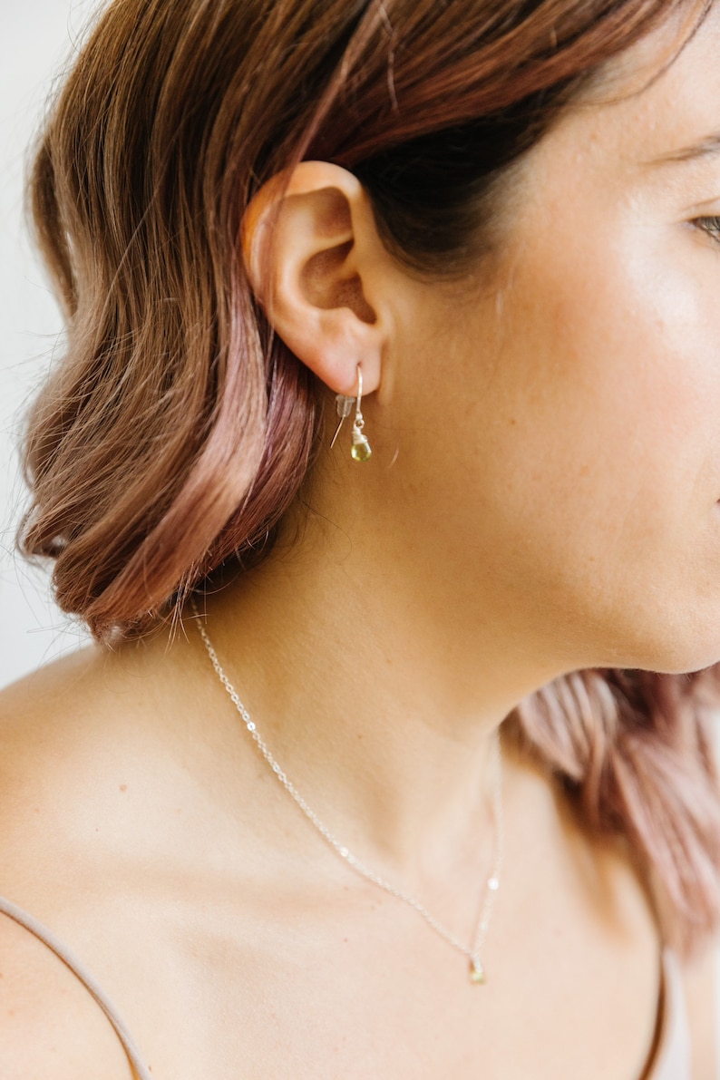 May include: A close-up of a woman wearing a silver necklace and earrings. The earrings are small, with a silver hook and a teardrop-shaped, light green gemstone. The necklace has a delicate chain with a matching gemstone pendant.