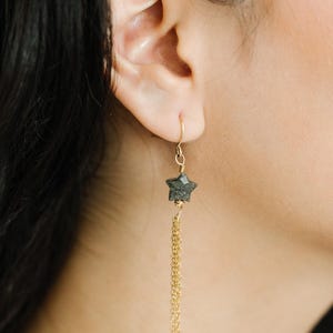 May include: Gold-tone star earrings with a dangling chain. The star-shaped charm is a dark, textured stone. The earrings are shown on a person's ear, with dark hair visible.