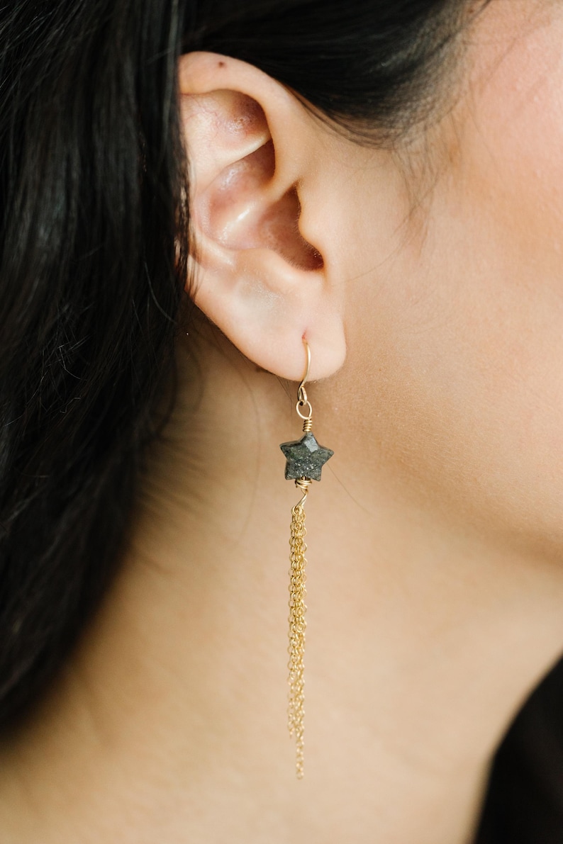 May include: Gold-tone star earrings with a dangling chain. The star-shaped charm is a dark, textured stone. The earrings are shown on a person's ear, with dark hair visible.