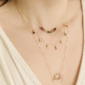 May include: A layered necklace set with three strands. The top strand features a curved row of colorful gemstones. The middle strand has small, dangling gemstones. The bottom strand has a large, clear gemstone pendant in a gold-tone setting.