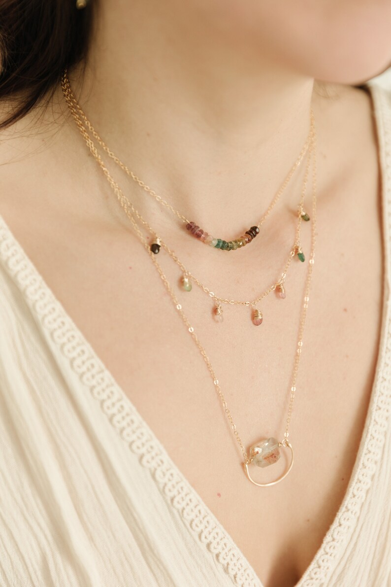 May include: A layered necklace set with three strands. The top strand features a curved row of colorful gemstones. The middle strand has small, dangling gemstones. The bottom strand has a large, clear gemstone pendant in a gold-tone setting.