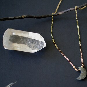 May include: A gold chain necklace with a crescent moon pendant. A clear quartz crystal and a dark twig are also in the image. The necklace is displayed on a dark background.